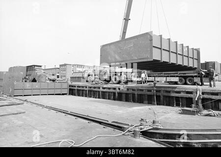 Transportinstallation fa. De Vries IJmuiden Transportunternehmen IJmuiden the Netherlands, Whizgle News, Dutch Desk, Niederlande, 1950 - 2000 am 12-07-1982. Dies sind die Elemente im Bild. Die Szene zeigt ein geschäftiges Industriegebiet mit Schwerpunkt auf schweren Maschinen und Arbeitern, die eine Aufgabe ausführen. Ein großer, metallischer Aufbau wird von einem Kran über einem Lkw gehoben, dessen starke Form einen Schatten auf den Boden wirft. Der Kran ist schräg positioniert, was seine Höhe und mechanische Komplexität unterstreicht. Im Vordergrund befindet sich eine Gruppe von Arbeitern in der Nähe des Lkws, der Stockfoto