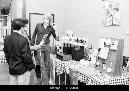 Holen Sie sich Ihr Gehör mit rechtem Stand Hörfachleute, Whizgle News, Dutch Desk, Niederlande, 1950 - 2000 im Jahr 1977. Die Abbildung zeigt diese Themen. Die Szene fängt ein lebhaftes Indoor-Event ein, möglicherweise eine Gemeinschaftsmesse oder eine Ausstellung. An vorderster Front führen drei Personen Gespräche, wobei ein Mann einen dunklen Anzug und eine Krawatte trägt, was auf ein formelles Verhalten hindeutet. Er steht mit gekreuzten Armen und hört einem anderen Teilnehmer zu, einem jüngeren Mann, der in einem lässigen, leicht aufgeknöpften Hemd gekleidet ist. Dieser Mann zeigt auf ein Array von Objekten, die auf einer Tabelle angezeigt werden, die verschiedene Merkmale aufweisen Stockfoto