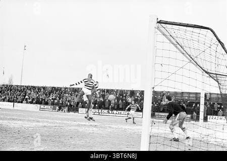 R.C.H. - Blue and White Football, Whizgle News, Dutch Desk, Niederlande, 1950 - 2000 am 03-02-1963. Diese Themen sind in der Abbildung dargestellt. In einer dynamischen Sportszene springt ein Spieler in einem gestreiften Trikot in die Luft und ist gekonnt bereit, ein entscheidendes Spiel auf dem Fußball zu machen. Der Spieler zeigt eine Mischung aus Agilität und Entschlossenheit, mit einem starken Fokus auf seinem Gesicht. Die markanten Streifen des Trikots stehen im Kontrast zu den gedämpften Tönen eines bewölkten Himmels und sorgen für einen auffälligen visuellen Effekt. Um den Spieler herum steht das Tor mit seinen weißen Pfosten, die das Geschehen einrahmen, deutlich sichtbar. Das Netting Stockfoto