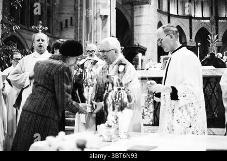 Weihe der Heiligölbasilika Haarlem Churches, Whizgle News, Dutch Desk, Niederlande, 1950 - 2000 am 04-04-1980. Diese Themen werden in der Abbildung angezeigt. In einem großen, aufwendig gestalteten Innenraum versammeln sich mehrere Individuen in einer feierlichen Atmosphäre, wahrscheinlich an einem Ort der Anbetung. Die Architektur verfügt über hohe, bogenförmige Fenster, die Licht eindringen lassen und weiche Schatten über die Steinoberflächen werfen. Die Menschen sind in formeller Kleidung gekleidet; einige tragen traditionelle Kleidung, komplett mit komplizierten Details, die auf einen zeremoniellen Anlass hinweisen. Im Vordergrund eine Frau in einem eleganten Muster Stockfoto
