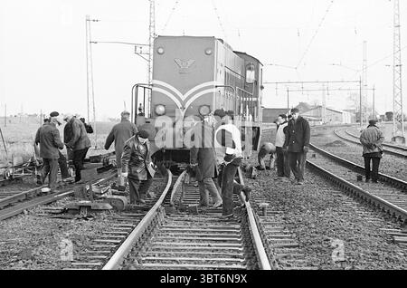 Lokomotive von der Schiene bei Halfweg Railways, Whizgle News, Dutch Desk, Niederlande, 1950 - 2000 am 16. 01. 1965. Die Abbildung zeigt diese Themen. Die Szene zeigt eine Eisenbahnanlage, die vor Aktivität strotzt, während eine Gruppe von Einzelpersonen auf den Gleisen arbeitet. Im Mittelpunkt des Bildes steht ein Zug mit einem markanten, kastenförmigen Motor, der in kräftigen dunkelgrünen und roten Farbtönen lackiert ist, was auf ein älteres Design hindeutet. Der Zug befindet sich auf den Gleisen, die aus einer Mischung aus Silber und rostigem Metall bestehen, was auf eine umfangreiche Nutzung über die Jahre hinweist. Rund um den Zug sind mehrere Arbeiter mit verschiedenen Aufgaben beschäftigt. Einige sind c Stockfoto