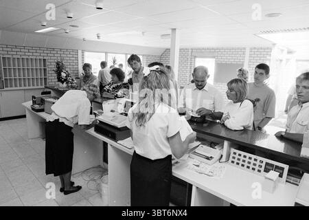 Vendorado Holiday Park Zandvoort: Holiday Park Reception Zandvoort, Whizgle News, Dutch Desk, Niederlande, 1950 - 2000 am 23-06-1989. Diese Themen sind in der Abbildung dargestellt. Die Szene zeigt einen belebten Innenbereich, wahrscheinlich einen Empfangsbereich oder einen Serviceschalter. Die dominante Farbpalette enthält verschiedene Graustufen, die das monochromatische Thema hervorheben. Die Wände sind meist hellgrau mit einer strukturierten Oberfläche, begleitet von einigen Ziegelsteinmauern, die das visuelle Interesse erhöhen. An der Vorderseite befindet sich ein Service-Schalter aus dunklem Material, der sich von der helleren Umgebung abhebt. Hinter den Zählern Stockfoto
