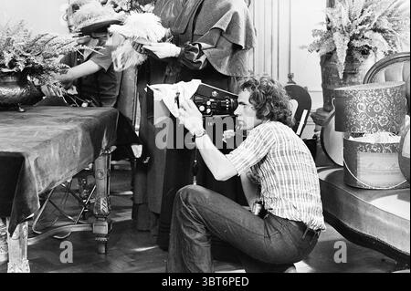 Film von Keetje Tippel mit Monique v.d. Ven. Dreharbeiten, Whizgle News, Dutch Desk, Niederlande, 1950 - 2000 am 23-08-1974. Dies sind die Themen im Bild. Die Szene spielt drinnen, geprägt von einer Vintage-Atmosphäre und etwas raffinierter Atmosphäre. Ein großer Tisch dominiert den Raum, bedeckt mit einem satten, dunklen Tuch, das einen Kontrast zu den hellen Tönen des darunter liegenden Holzbodens bildet. Mehrere Topfpflanzen mit üppigem, grünem Laub sind um den Raum herum angeordnet und verleihen der Umgebung einen Hauch von Leben und Farbe. Im Vordergrund hockt sich ein Mann hinter einem klassischen Kameraaufbau, equ Stockfoto