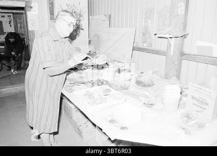 Mrs. Gordijn mit altem Weinfass IJmuiden Archeology, Whizgle News, Dutch Desk, Niederlande, 1950 - 2000 am 08-01-1977. Das Bild enthält diese Themen. Die Szene fängt ein rustikales Interieur ein, wahrscheinlich in einem Gemeindezentrum oder einer Messe. Eine Frau in einem Vintage-gestreiften Kleid steht an einem Tisch und konzentriert sich dabei auf eine Auswahl von Gegenständen, die vor ihr liegen. Ihre Brille reflektiert ein sanftes Licht, was zu einem fleißigen und anspruchsvollen Look beiträgt, während sie die Ausstellungsstücke inspiziert. Das Ambiente ist warm und einladend, was ein Gefühl von Gemeinschaft und gemeinsamen Interessen suggeriert. Der Tisch ist mit verschiedenen Kontai geschmückt Stockfoto