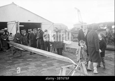 Bootstaufe 'de Stern' IJmuiden' Ruderwettbewerbe etc., Whizgle News, Dutch Desk, Niederlande, 1950 - 2000 am 02-01-1965. Diese Themen werden in der Abbildung angezeigt. In der Szene versammelt sich eine Gruppe von Menschen im Freien neben einem großen, offenen Gebäude, das einer Werkstatt oder einem Schuppen ähnelt. Der Boden ist mit rechteckigen Steinen gepflastert, was zu einer rustikalen und doch organisierten Atmosphäre beiträgt. Es gibt ein langes Holzboot, teilweise fertiggestellt, auf Sägetieren ruht, das sich über den Vorderboden erstreckt. Die Menge besteht aus Erwachsenen und Kindern, die meist in dunkler und gedämpfter Kleidung gekleidet sind. Stockfoto