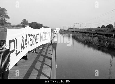 Banner auf Brücke Haarlemmerliede Banners Haarlemmerliede, Whizgle News, Dutch Desk, Niederlande, 1950 - 2000 am 28-09-1984. Diese Themen sind in der Abbildung dargestellt. Die Szene fängt einen Moment des Aktivismus an einem hölzernen Pier ein, der sich über ein ruhiges Gewässer erstreckt. Auf der einen Seite steht eine Gruppe von Menschen, die große Banner halten, die den Vordergrund dominieren. Diese Banner sind stark gegen die gedämpften Töne der Umgebung und unterstreichen ihre Botschaft. Die Individuen tragen lässige Kleidung, die ein Gefühl der Verbundenheit verleiht, und ihre Haltung vermittelt eine Mischung aus Entschlossenheit und Hoffnung. Die Stockfoto