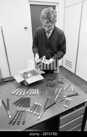 Konfiszierte Schmetterlingsmesser in der Haarlem Polizeistation Haarlem, Niederlande, Whizgle News, Dutch Desk, Niederlande, 1950 - 2000 am 03-01-1989. Die Abbildung zeigt diese Themen. In einem leicht beleuchteten Innenraum steht ein junger Mann über einem Holztisch und nimmt eine Aufgabe wahr, die sorgfältige Aufmerksamkeit erfordert. Er trägt einen dunklen, lässigen Pullover, der über einem Hemd mit Kragen getragen wird, und seine Haare sind leicht zerzaust, was auf eine entspannte Atmosphäre hindeutet. Der Tisch ist überfüllt mit einer Reihe von Objekten – mehrere ordentlich angeordnete Stapel von kleinen, rechteckigen Gegenständen, wahrscheinlichen Werkzeugen oder Instrumenten, Stockfoto