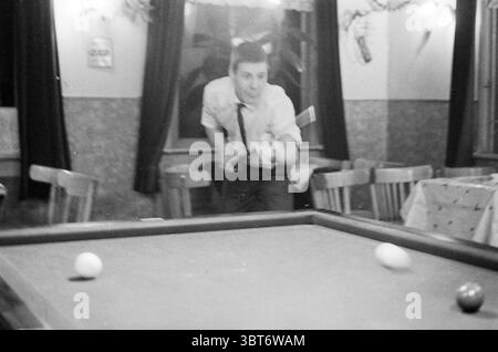 Family de Boer im Café Brabant Café, Whizgle News, Dutch Desk, Niederlande, 1950 - 2000 am 25-12-1960. Dies sind die Elemente im Bild. Die Szene spielt in einem gemütlichen, schwach beleuchteten Raum, der einen Vintage-Charme ausstrahlt, wahrscheinlich eine Taverne oder ein Erholungsraum. Die Wände sind mit dezenten Dekorationen verziert, die auf frühere Feste oder Zusammenkünfte hinweisen. Sanfte, gedämpfte Töne dominieren den Raum, warme Brauntöne und sanfte Cremes betonen die Atmosphäre. Im Vordergrund steht ein Mann an einem Billardtisch und greift mit konzentrierter Entschlossenheit einen Queustock. Er trägt ein weißes Hemd, das cris kontrastiert Stockfoto