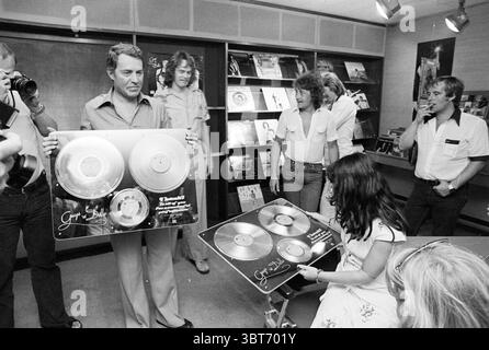Gold-Album Guys and Dolls Negram Music, Whizgle News, Dutch Desk, Niederlande, 1950 - 2000 am 06-07-1977. Das Bild enthält diese Themen. In einem lebendigen Innenbereich versammelt sich eine Gruppe von Menschen um zwei zentrale Figuren. Eine Person trägt ein helles Hemd und eine gut geschnittene Hose und hält zwei große, gerahmte Goldplatten, die ihre glänzenden Metallic-Oberflächen deutlich sichtbar zeigen. Ihr Ausdruck strahlt Stolz und Freude aus, wenn sie mit der Gruppe interagieren. Neben ihnen untersucht eine sitzende Person, eine Frau mit dunklen Haaren, eine andere Reihe von Aufzeichnungen vor ihr, zeigt einen Blick von Aufregung und Stockfoto