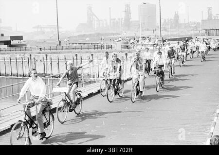 Christel Adelaar auf Fahrrad Haarlem-Velsen Radwege Radfahrer, Whizgle News, Dutch Desk, Niederlande, 1950 - 2000 am 08-06-1963. Das Bild enthält diese Themen. Die Szene zeigt eine lebhafte Gruppe von Radfahrern, die eine hölzerne Promenade entlang eines großen Wassers durchquert. Eine Vielzahl von Menschen wird beim Pedalieren beobachtet, die verschiedene Haltungen und Stile zeigen. Einige Fahrer winken fröhlich und verkörpern ein Gefühl von Kameradschaft und Vergnügen. Im Vordergrund fährt ein Mann links mit dem Fahrrad, während er ein helles Hemd trägt und ein lässiges Verhalten ausstrahlt. Direkt neben ihm, ein temperamentvolles W Stockfoto