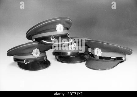 Polizeisprache, Whizgle News, Dutch Desk, Niederlande, 1950 - 2000 am 05-05-1991. Das Bild enthält diese Themen. Die Szene zeigt eine Sammlung von vier Polizeihüten, die in einer mehrschichtigen Formation angeordnet sind. Jeder Hut zeichnet sich durch sein unverwechselbares Design aus: Dunkler Stoff mit einem glänzenden schwarzen Visier und einem glänzenden Emblem, das auf der Vorderseite sichtbar ist. Die Hüte werden vor einem sanft beleuchteten Hintergrund platziert, der von hellgrau zu dunkleren Schattierungen wechselt und eine monochromatische Atmosphäre schafft. Die Hüte variieren leicht in Neigung und Ausrichtung, was zu einem Gefühl von Tiefe und Dimension beiträgt. Das Licht fängt ein Stockfoto