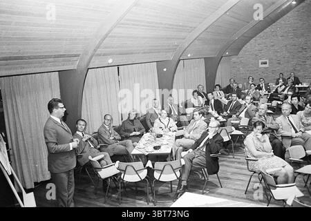 Heemskerk Meetings, Whizgle News, Dutch Desk, Niederlande, 1950 - 2000 am 20-08-1969. Das Bild enthält diese Themen. Die Szene präsentiert ein Treffen in einem geräumigen Innenraum, das sich durch sein einzigartiges architektonisches Design mit hohen, geschwungenen Holzdecken auszeichnet, die ein Gefühl von Wärme und Offenheit schaffen. Die Wände sind mit einer gedämpften Backsteinstruktur verziert, die der Umgebung ein gemütliches und zugleich formelles Ambiente verleiht. Im Vordergrund steht ein Mann mit Gesten, während er ein sitzendes Publikum anspricht. Er trägt einen formellen Anzug mit Brille, der ein Gefühl von Autorität und enga ausstrahlt Stockfoto