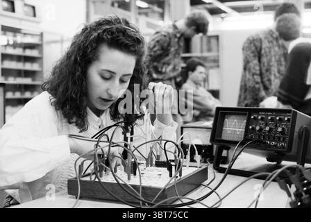 Matty Snoep MTS Santpoort Santpoort, Whizgle News, Dutch Desk, Niederlande, 1950 - 2000 am 12-03-1991. Diese Themen sind in der Abbildung dargestellt. Die Szene fängt eine geschäftige Innenumgebung ein, wahrscheinlich ein Labor oder ein Klassenzimmer. Im Mittelpunkt der Komposition steht eine junge Frau mit lockigem Haar, die sich intensiv auf eine komplexe Palette von Geräten konzentriert, die vor ihr liegen. Sie trägt eine weiße, strukturierte Bluse, die einen starken Kontrast zu den dunkleren Farben der Umgebung bildet. Vor ihr befindet sich eine organisierte Holztafel mit verschiedenen elektronischen Bauteilen, von denen zahlreiche bunte Drähte hinausgehen. Die Stra Stockfoto