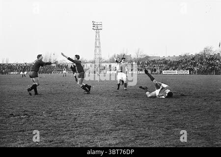 Haarlem - H.V.C. Football, Whizgle News, Dutch Desk, Niederlande, 1950 - 2000 am 01-12-1963. Die Abbildung zeigt diese Themen. Die Szene fängt einen intensiven Moment eines Fußballspiels in einem grasbewachsenen Stadion voller Zuschauer ein. Im Vordergrund wird ein Spieler in einem weißen Trikot mit hochgezogenen Beinen auf dem Boden gesehen, was darauf hindeutet, dass er während des Spiels umgeworfen wurde. Der Ausdruck auf seinem Gesicht vermittelt eine Mischung aus Frustration und Entschlossenheit. Um ihn herum befinden sich Spieler in kontrastierenden dunklen Trikots, von denen einer scheinbar mit ausgestreckten Armen feiert, was auf ein jüngstes Tor oder eine bedeutende Bedeutung hinweist Stockfoto