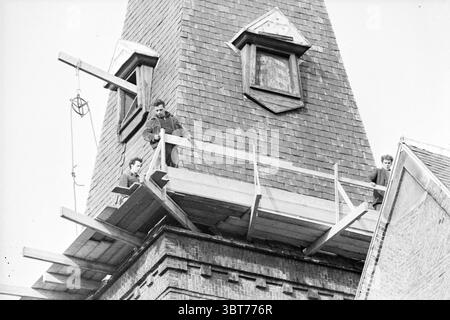 Restaurierung der Wilhelminenkirche GED.Oude Gracht Kirchen außer römisch-katholischen. Haarlem Gedempte Oude Gracht the Netherlands, Whizgle News, Dutch Desk, Niederlande, 1950 - 2000 am 14-03-1963. Dies sind die Elemente im Bild. In einer Szene, die von einem rustikalen Gebäude dominiert wird, sitzen drei Individuen auf einem Holzgerüst, das aus einem oberen Stockwerk eines hohen Gebäudes ragt. Die Architektur verfügt über ein steiles, spitzes Dach und unterschiedliche Giebelabschnitte, die dem Äußeren Charakter verleihen. Die Oberfläche besteht aus roten und braunen Fliesen, die zu einem historischen Flair beitragen. Eine Zahl ist sli Stockfoto