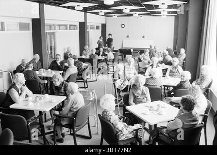 Altersheim, Whizgle News, Dutch Desk, Niederlande, 1950 - 2000 auf 01-1985. Die Abbildung zeigt diese Themen. Die Szene zeigt einen geräumigen Innenbereich mit einer großen Gruppe älterer Personen, die an runden Tischen sitzen. Die Komposition zeigt eine Mischung aus Interaktionen, wobei einige Menschen lebhafte Gespräche führen, während andere scheinbar leise beobachten oder einen Moment der Reflexion genießen. Die Farbpalette ist überwiegend gedämpft und bevorzugt Grautöne und sanfte Weißtöne, was dem Raum eine nostalgische oder zeitlose Atmosphäre verleiht. Die Wände sind in einer hellen Farbe gestrichen, die einen Kontrast zum schwarzen Elem bildet Stockfoto