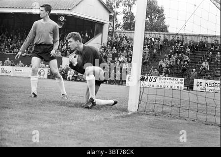 RCH - EDO Football, Whizgle News, Dutch Desk, Niederlande, 1950 - 2000 am 25-08-1963. Das Bild enthält diese Themen. Die Szene fängt einen Moment in einem Fußballspiel ein, vor der Kulisse eines belebten Stadions voller Zuschauer, die das Spiel mit Spannung beobachten. Der Rasen ist üppig, leuchtend grün, während die Linien, die das Feld markieren, klar und klar sind, was zu einem Gefühl der Begeisterung beiträgt. Im Vordergrund befinden sich zwei Spieler in der Nähe eines der Torpfosten. Eine, die in einem langärmeligen grauen Hemd gekleidet ist, scheint aufmerksam zu sein und bereitet sich wahrscheinlich auf die Handlung vor. Seine Haltung deutet auf Konzentration und Abschreckung hin Stockfoto