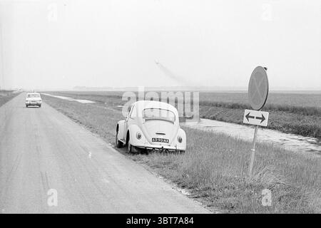 Stoppen Sie Ban Vijfhuizerweg. Spotspot für Flugzeuge..., Whizgle News, Dutch Desk, Niederlande, 1950 - 2000. Die Abbildung zeigt diese Themen. Die Szene fängt eine ruhige, offene Landschaft ein, die überwiegend in sanften Grautönen gefärbt ist, was auf einen bewölkten Himmel hindeutet. Neben einer geraden, leeren Straße ruht ein Oldtimer, der sich durch seine abgerundete Form und sein unverwechselbares Design auszeichnet, teilweise auf üppigem grünem Gras vom Bürgersteig ab. Seine helle Farbe steht in zartem Kontrast zu den umgebenden Erdtönen. Auf der anderen Straßenseite ist ein anderes Fahrzeug sichtbar, das in einer Entfernung geparkt ist und dem ein Hauch von Leben verleiht Stockfoto