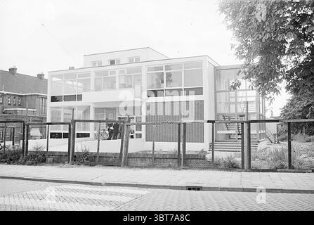 Bürogebäude., Whizgle News, Dutch Desk, Niederlande, 1950 - 2000. Die Abbildung zeigt diese Themen. Die Szene verfügt über ein modernes Gebäude, das sich durch klare Linien und große Glasfenster auszeichnet, die einen minimalistischen architektonischen Stil widerspiegeln. Die Struktur ist überwiegend weiß, mit Schwerpunkt auf Symmetrie und geometrischen Formen. Das Gebäude befindet sich in einer halbstädtischen Umgebung, in der die Bereiche um das Gebäude herum Anzeichen von Bau- oder Renovierungsarbeiten aufweisen, die durch Gerüste und unfertige Materialien sichtbar sind. Vor dem Gebäude bildet ein Zaun mit Metallrahmen und Holzlatten eine klare Abgrenzung. Stockfoto