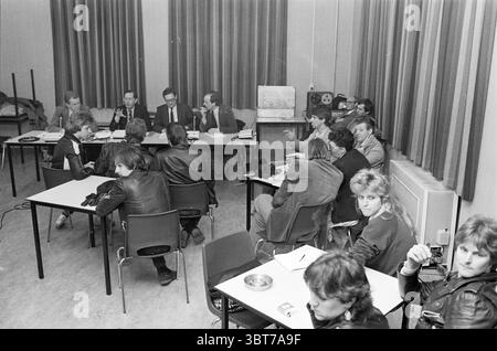 Information Evening, Whizgle News, Dutch Desk, Niederlande, 1950 - 2000 am 03-1983. Dies sind die Elemente im Bild. Die Szene zeigt eine Zusammenkunft in einem Klassenzimmer oder Besprechungsraum, die sich Retro anfühlt, wahrscheinlich aus dem späten 20. Jahrhundert. Der Raum ist voll mit Einzelpersonen, hauptsächlich jungen Erwachsenen, die um mehrere Tische sitzen. Das Layout besteht aus zwei prominenten Gruppen: Eine besteht aus einer Handvoll Einzelpersonen in Anzügen, die an der Vorderseite sitzen und wahrscheinlich an Diskussionen oder Präsentationen teilnehmen, während die andere Gruppe aus Studenten oder Teilnehmern besteht, die an den Tischen verstreut sind und das Verfahren beobachten Stockfoto