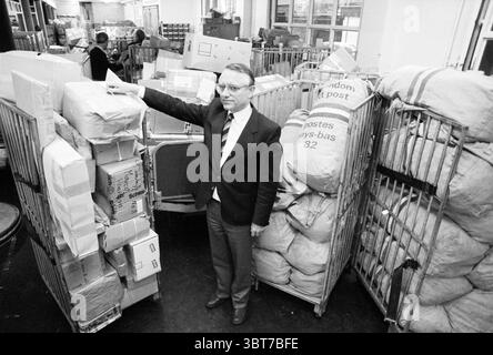 Direktor den Boer Haarlem Zwischen Bergen der Post PTT Postdienste City Mail Haarlem the Netherlands, Whizgle News, Dutch Desk, Niederlande, 1950 - 2000 am 28-11-1983. Diese Themen werden in der Abbildung angezeigt. Die Szene zeigt einen belebten Postsortierbereich mit gestapelten Kartons und Taschen. Im Vordergrund steht ein Mann in dunklem Anzug und Krawatte selbstbewusst neben einem großen Stapel von Paketboxen, von denen viele in braunes Papier gewickelt und mit Schnur gebunden sind. Seine Haltung ist entspannt, aber autoritativ, da er eine Hand auf eine Box legt und direkt auf den Betrachter blickt. Um ihn herum, die Stockfoto