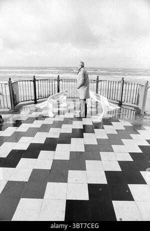 The Roundabout' Zandvoort Zandvoort Strandweg, Whizgle News, Dutch Desk, Niederlande, 1950 - 2000 am 12-11-1991. Das Bild enthält diese Themen. In einer Küstenszene steht eine Einzelfigur auf einer gemusterten Fliesenfläche mit einem Schachbrettmuster aus schwarzen und weißen Quadraten. Der Mann, in einem hellen Mantel und einer Fedora gekleidet, sieht sich der Weite des Ozeans gegenüber, scheinbar verloren in Gedanken. Der Horizont verschmilzt in einen Himmel voller gedämpfter grauer Wolken, die auf einen bewölkten Tag hindeuten. Die Wellen des Ozeans schlagen sanft gegen das Ufer, ihre Bewegungen erzeugen einen rhythmischen Klang, der sich ergänzen kann Stockfoto
