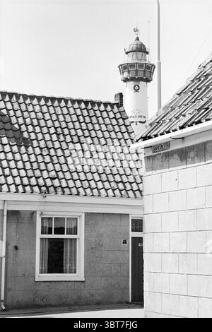 Bericht Egmond aan Zee: Prins Hendrik Foundation Oude Dorpsplein Egmond aan Zee Egmond aan Zee Oude Dorpsplein, Whizgle News, Dutch Desk, Niederlande, 1950 - 2000 am 23-05-1977. Dies sind die Elemente im Bild. Die Szene fängt eine ruhige urbane Umgebung ein, eingerahmt von einer Mischung aus architektonischen Elementen. Im Vordergrund bildet eine niedrige Mauer aus hellem Stein eine Kulisse, daneben steht ein traditionelles Gebäude mit einem schrägen Dach aus dunkelroten Fliesen. Die Dachziegel sind in einem willkürlichen Muster angeordnet, das Alter und Charakter angibt. Hinter diesen Gebäuden erhebt sich ein hoher Leuchtturm Stockfoto