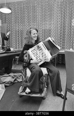 Handing over Musikinstrumente Heliomare Wijk aan Zee Handover and Transfers, Whizgle News, Dutch Desk, Niederlande, 1950 - 2000 am 11-01-1977. Das Bild enthält diese Themen. In einem Zimmer mit einer warmen und einladenden Atmosphäre sitzt eine Frau im Rollstuhl und hält ein Akkordeon. Ihr Haar ist lang und lockig, was ihr eine lebendige Präsenz verleiht. Die Akkordeon ist groß und verfügt über eine Reihe von schwarzen und weißen Tasten, die sich im Kontrast zu den insgesamt warmen Tönen in der Umgebung befinden. Der Hintergrund zeigt einen gemusterten Vorhang, der eine subtile Textur mit geometrischen Formen in gedeckten Farben verleiht Stockfoto