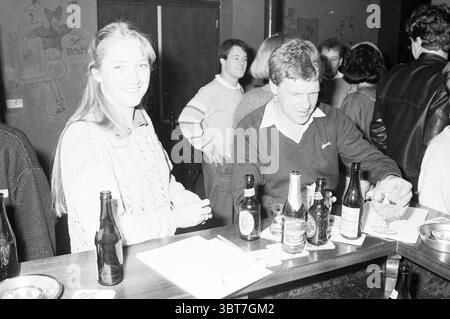 Bierverkostungstest Nieuw-Vennep Getränke- und Spirituosengeschäfte Nieuw-Vennep, Whizgle News, Dutch Desk, Niederlande, 1950 - 2000 am 11-11-1984. Dies sind die Themen im Bild. Die Szene fängt eine lebhafte Bar-Atmosphäre mit einer Mischung von Gästen ein, die sich unterhalten und ihre Drinks genießen. Im Vordergrund steht eine Frau mit einem warmen Lächeln und trägt einen hellen Strickpullover. Ihr Haar ist lang und leicht zerzaust, was das dunkle Licht der Einrichtung einfängt. Neben ihr steht ein Mann, der sich darauf konzentriert, ein Getränk aus einer Flasche in ein Glas zu gießen. Er trägt ein dunkles Hemd mit Kragen und ein sportliches Hemd Stockfoto