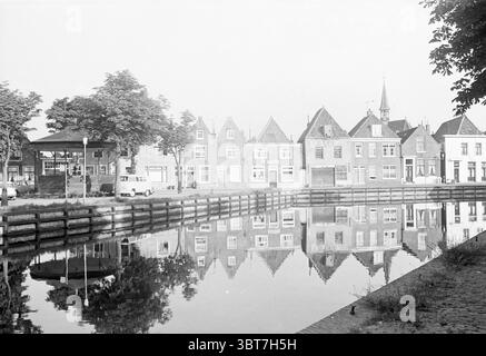 Blick auf die Kammer Spaarndam Westkolk., Whizgle News, Dutch Desk, Niederlande, 1950 - 2000. Die Abbildung zeigt diese Themen. Die Szene zeigt einen ruhigen Kanal, der von malerischen, historischen Gebäuden umgeben ist, von denen jedes von schrägen Dächern und bezaubernden Fassaden geprägt ist. Die Strukturen sind überwiegend in sanften, gedämpften Tönen dargestellt, die Grautöne, Weiße und Anklänge von Erdfarben vermischen, was der Gesamtkomposition ein nostalgisches Gefühl verleiht. Hohe Bäume säumen den Kanal, ihre üppig grünen Blätter bilden ein sanftes Baldachin, das den Boden mit Licht- und Schattenflecken bedeckt. Die Oberfläche des Wassers ist spiegelförmig, Stockfoto
