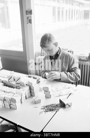 Sozialer Workshop (bei Meer en Bosch?) Jugendliche und Erwachsene packen Boxen mit Schrauben, Whizgle News, Dutch Desk, Niederlande, 1950 - 2000 im Jahr 1963. Dies sind die Elemente im Bild. In einem gut beleuchteten Innenbereich ist ein Junge intensiv an einer Aktivität an einem Tisch beteiligt. Er trägt einen hellen Mantel, der im Kontrast zu den dunkleren Tönen der Tischoberfläche steht. Die Haltung des Jungen ist fokussiert; er neigt sich leicht nach vorne mit dem Kopf nach unten und nimmt sich in die anstehende Aufgabe auf. Um ihn herum sind verschiedene Gegenstände ordentlich angeordnet. Eine große Sammlung von kleinen rechteckigen Kästen ist auf dem Tisch gestapelt, was auf einen hindeutet Stockfoto