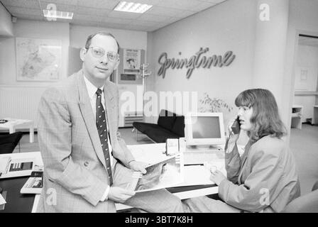 Interior Springtime, Whizgle News, Dutch Desk, Niederlande, 1950 - 2000 am 29-08-1991. Das Bild enthält diese Themen. In einer modernen Büroumgebung sind zwei Personen in ihrer Arbeitsumgebung engagiert. Links hält ein Mann in einem hellgrauen Anzug eine illustrierte Broschüre oder ein Dokument, die eine Nahaufnahme einer malerischen Landschaft zeigt. Sein Verhalten ist selbstbewusst, mit einem leichten Lächeln, und er blickt direkt auf den Zuschauer. Seine Brille reflektiert dezentes Licht, und er trägt eine Krawatte mit einem Muster aus kleinen Punkten, die seiner formellen Kleidung einen Hauch von Persönlichkeit verleiht. Rechts sitzt eine Frau an einem Schreibtisch. Stockfoto