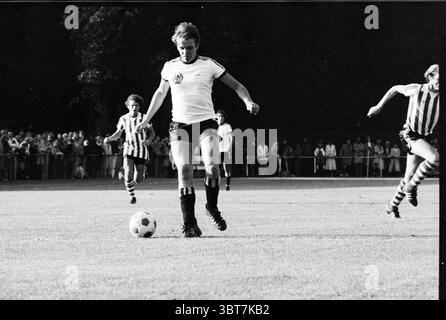 Fußball: HFC - PSV Football, Whizgle News, Dutch Desk, Niederlande, 1950 - 2000 am 14-08-1979. Das Bild enthält diese Themen. Die Szene fängt einen dynamischen Moment auf einem grasbewachsenen Feld ein, gefüllt mit der Energie eines Fußballspiels. An der Spitze befindet sich ein fokussierter Spieler in einem strahlend weißen Trikot mit schwarzem Besatz im mittleren Schritt, der einen Fußball präzise nach vorne treibt. Sein sportlicher Aufbau deutet auf Intensität und Entschlossenheit hin. Die Shorts, die er trägt, sind ebenfalls schwarz und ergänzen das gesamte monochrome Thema der Kleidung. Im Hintergrund ist ein kontrastierendes Streifenmuster auf einem anderen Spieler sichtbar Stockfoto
