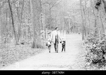 Herr Heemskerk in de Hout Personen, Whizgle News, Dutch Desk, Niederlande, 1950 - 2000 am 10-03-1972. Die Abbildung zeigt diese Themen. In einer ruhigen Waldlandschaft spaziert ein Mann auf einem schmalen, von hohen Bäumen flankierten Pfad, deren Stämme lange Schatten im weichen, diffusen Licht werfen. Die Atmosphäre ist ruhig und ruhig, was auf eine sanfte Stimmung hindeutet. Das Laub besteht überwiegend aus gedämpftem Grün, Anklängen von Grau und Erdbräunen, die die natürliche Umgebung betonen. Der Mann trägt einen hellen Trenchcoat, der im Kontrast zu den dunkleren Tönen des Waldes steht. Er hält Hände mit zwei Kindern, die Stockfoto