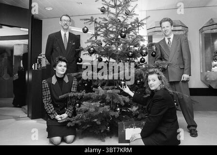 Juwelier Aronson Christmas Tree, Whizgle News, Dutch Desk, Niederlande, 1950 - 2000 am 08-12-1991. Das Bild enthält diese Themen. In gemütlicher Innenumgebung steht ein gut dekorierter Weihnachtsbaum im Mittelpunkt, dessen Zweige mit glänzenden schwarzen Ornamenten beladen sind, die das Licht reflektieren. Um den Baum herum befinden sich vier Individuen, die in einer freundlichen Gruppe stehen. Links steht ein Mann in einem Anzug mit Brille, der ein formelles, aber dennoch zugängliches Verhalten ausstrahlt. Seine Kleidung, ein klassischer dunkler Anzug mit weißem Hemd, ergänzt die Urlaubsatmosphäre. Rechts ein anderer Mann in einem ähnlichen Anzug, aber leicht Stockfoto