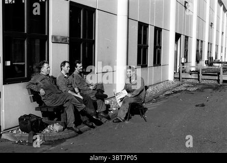 Vier Männer machen eine Pause. Whizgle News, Dutch Desk, Niederlande, 1950 - 2000. Diese Themen sind in der Abbildung dargestellt. In einer schlichten, industriellen Umgebung sitzen vier Personen auf einer Bank neben einem modernen Gebäude, das durch große rechteckige Fenster gekennzeichnet ist, die das Licht reflektieren. Die Architektur wirkt utilitaristisch, mit einer Fassade, die eine Mischung aus gedämpften Farben zeigt – Grau, weiß und dezenten Blautönen. Die Menschen, die in Arbeitskleidung gekleidet sind, die einen praktischen Zweck nahelegt, strahlen ein entspanntes Verhalten aus. Ihre Kleidung ist überwiegend dunkel und fügt sich in die düsteren Töne ihrer Umgebung ein. Stockfoto