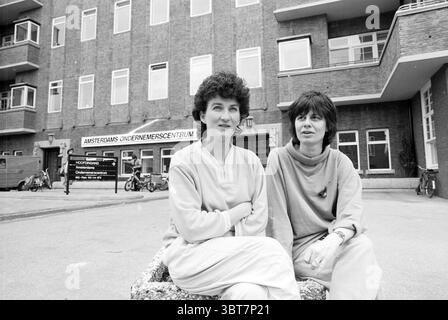 Women and IT Bureau Amsterdam People Companies Women Amsterdam the Netherlands, Whizgle News, Dutch Desk, Niederlande, 1950 - 2000 am 26-03-1987. Das Bild enthält diese Themen. In einer geschäftigen städtischen Umgebung sitzen zwei Frauen auf einem Steinbau zusammen und wirken entspannt und in Gesprächen verwickelt. Die Frau auf der linken Seite hat lockiges, dunkles Haar und trägt ein helles, lässiges Oberteil, während die Frau auf der rechten Seite kurze, glatte Haare hat und ein etwas dunkleres, übergroßes Kleidungsstück trägt. Der Hintergrund ist ein markantes Backsteingebäude mit großen Fenstern und modernem Design. Das Archit Stockfoto