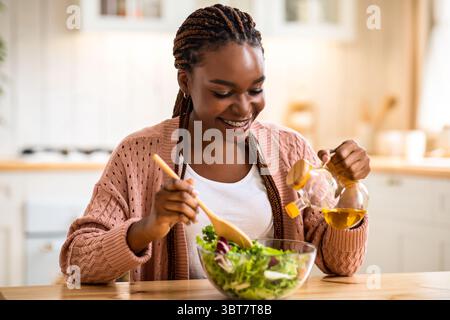 Lächelnde afroamerikanische Frau kocht Gemüsesalat in der Küche und fügt Olivenöl hinzu Stockfoto