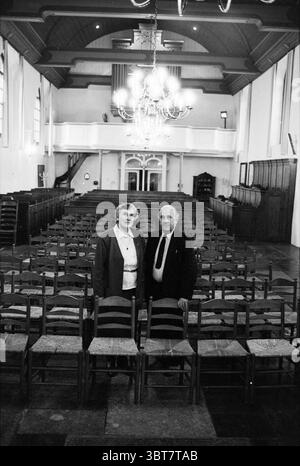 Van Riessen sexton Engelmunduskerk IJmuiden IJmuiden the Netherlands, Whizgle News, Dutch Desk, Niederlande, 1950 - 2000 am 07-01-1991. Dies sind die Elemente im Bild. Die Szene zeigt einen weitläufigen, leeren Saal, der durch Reihen von Holzstühlen gekennzeichnet ist, die ordentlich in einem gitterartigen Muster angeordnet sind. An vorderster Front steht ein älteres Paar zusammen und strahlt ein Gefühl von Wärme und Gesellschaft aus. Die Frau trägt einen taillierten Blazer über einer hellen Bluse, während der Mann einen formellen Anzug und Krawatte trägt, was ein würdevolles Aussehen verleiht. Die Architektur der Halle hat hohe Decken, die mit wi verziert sind Stockfoto