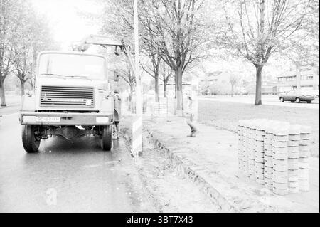 Ersetzung von Grünanlagen für Beton Heerenduinweg Works IJmuiden Heerenduinweg the Netherlands, Whizgle News, Dutch Desk, Niederlande, 1950 - 2000 am 21. 10. 1986. Dies sind die Elemente im Bild. In einer Szene, die von verschiedenen Grautönen dominiert wird, wird ein großes Baufahrzeug auf einer leicht nassen Straße deutlich geparkt. Der Lkw hat ein robustes Erscheinungsbild mit robusten Rädern und einer großen Kabine. Ein Arbeiter, der in praktischer Kleidung gekleidet ist, scheint in der Nähe des Fahrzeugs an einer Aufgabe beteiligt zu sein und zeigt ein fokussiertes Verhalten während der Bautätigkeiten. Rechts, ordentlich gestapelte Steinblöcke Stockfoto