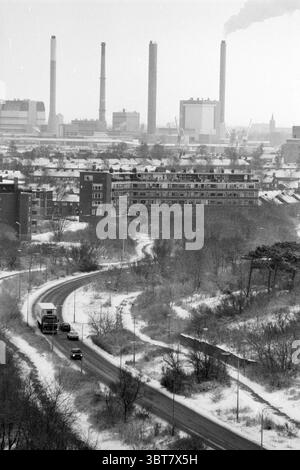 Zeewijk IJmuiden Snow IJmuiden the Netherlands, Whizgle News, Dutch Desk, Niederlande, 1950 - 2000 am 13-02-1991. Die Abbildung zeigt diese Themen. Die Szene zeigt eine schroffe Industrielandschaft, die von der Dominanz von Schornsteinen geprägt ist, die sich gegen einen gedämpften, bewölkten Himmel erheben. Verschiedene hohe Schornsteine sind sichtbar, was zu einem Gefühl von Schwere und Dunkelheit beiträgt. Im Vordergrund schlängelt sich eine kurvige Straße durch das Gelände, gesäumt von minimalem Laub, was auf eine Winterkulisse mit leichtem Schnee hinweist. Auf der einen Seite befinden sich mehrere flache Gebäude, deren Backsteinfassaden sich in die einfügt Stockfoto