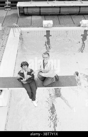 Schwimmbad de Vosse: Viertägiges Schwimmen, Whizgle News, Dutch Desk, Niederlande, 1950 - 2000 am 02-06-1991. Die Abbildung zeigt diese Themen. In einer ruhigen Umgebung im Freien spiegelt der Swimmingpool eine ruhige Atmosphäre wider, seine Oberfläche glitzert unter sanfter Beleuchtung. Der Pool ist von einer Betonterrasse umgeben, und mehrere Sprungbretter erstrecken sich über das Wasser. Im Vordergrund sitzen eine Frau und ein Mann am Rand eines Sprungbretts. Die Frau, die eine sportliche Jacke trägt, wirkt besinnlich, mit den Händen auf den Knien. Ihr Haar ist lässig gestylt, was zu einer entspannten Atmosphäre beiträgt. Zu ihrem Outfit gehören Stockfoto