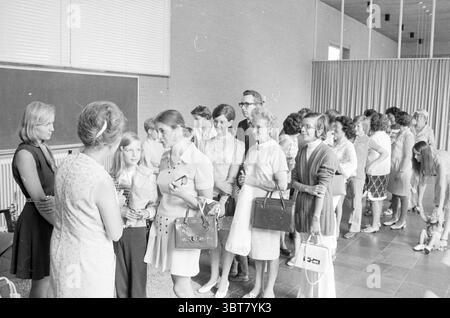 Kinder gratulieren Lehrer (Abschied/Jubiläum), Whizgle News, Dutch Desk, Niederlande, 1950 - 2000 auf 07-1971. Die Abbildung zeigt diese Themen. Die Szene zeigt eine Gruppe von Menschen, vor allem Frauen, die sich in einem geräumigen Innenraum versammelt haben, der sich durch eine minimalistische Ästhetik auszeichnet. Die Wände sind hell, möglicherweise weiß oder beige, was eine helle und luftige Atmosphäre schafft. Große Fenster lassen natürliches Licht den Bereich durchfluten, beleuchten die Gesichter der Menschen und verstärken das Gefühl der Offenheit. Im Vordergrund steht eine Frau mit ihrem langen, hellen Haar, das einen Ärmel trägt Stockfoto