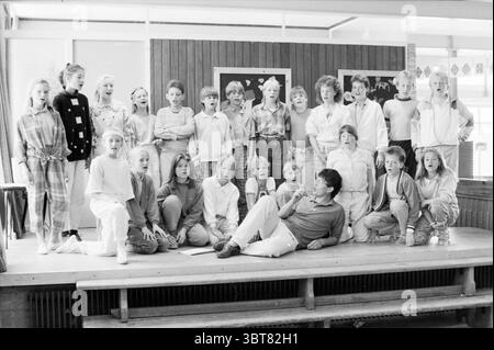 Probe Musical Pleiadesschool-Probe, Whizgle News, Dutch Desk, Niederlande, 1950 - 2000 auf 30-05-1986. Das Bild enthält diese Themen. Die Szene zeigt eine große Gruppe von Kindern und einen Erwachsenen, die sich auf einer Holzbühne versammeln. Die Bühne befindet sich in einem hellen Raum, der von natürlichem Licht beleuchtet wird, das durch große Fenster hereinströmt. Die Wände sind mit Holztäfelungen verziert, die eine warme Kulisse schaffen, vor der die Figuren stehen. Die Kinder, Jungen und Mädchen, tragen eine Mischung aus lässiger Kleidung, hauptsächlich in hellen und pastellfarbenen Farben. Einige tragen gemusterte Outfits, andere tragen Stockfoto