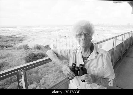 Mrs. Van de Berg schaut sich die Segelschiffe in IJmuiden von ihrer Heimat IJmuiden in den Niederlanden an. Whizgle News, Dutch Desk, Niederlande, 1950 - 2000. Das Bild enthält diese Themen. Die Szene zeigt eine ältere Frau, die auf einem Balkon steht, umgeben von einem weiten Blick auf eine Stadtlandschaft mit industriellen und urbanen Elementen. Ihr weißes Haar, flauschig und voluminös, steht im Kontrast zu den dunkleren Tönen eines hellgewaschenen Hemdes, das sie trägt. Sie hält ein Fernglas in der Hand und hält es fest, als ob es ein wesentlicher Teil ihrer Erkundung wäre. Der Hintergrund zeigt eine Wucherung Stockfoto