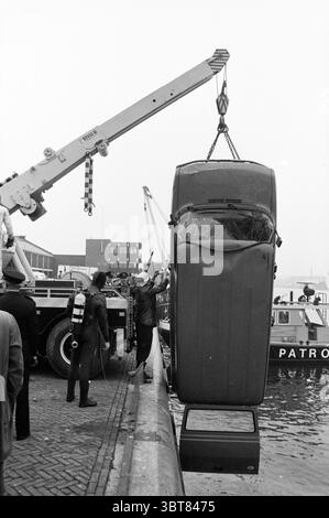 Polizeiwagen im Wasser IJmuiden Police IJmuiden the Netherlands, Whizgle News, Dutch Desk, Niederlande, 1950 - 2000 am 14-05-1985. Das Bild enthält diese Themen. Die Szene spielt in einem Hafen, der durch eine nebelige Atmosphäre gekennzeichnet ist, die das natürliche Licht subtil streut und eine düstere und gedämpfte Atmosphäre schafft. Ein großer Kran, der in Industriestruktur gelb und schwarz lackiert ist, dominiert die linke Seite, der seinen Arm über das Wasser streckt. Der Haken des Krans ist fest an der Oberseite eines Oldtimers befestigt, der vertikal angehoben wird; seine Räder sind nach unten gerichtet, und der Wagen scheint ein klassischer Mod zu sein Stockfoto