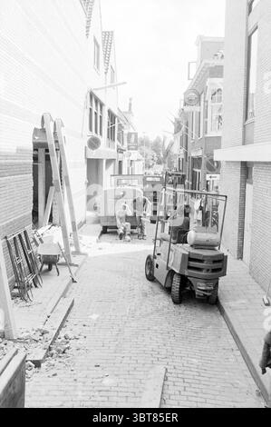 Nieuwstraat B'wijk Straten Beverwijk Nieuwstraat Niederlande, Whizgle News, Dutch Desk, Niederlande, 1950 - 2000 auf 18-08-1978. Dies sind die Elemente im Bild. In einer engen, belebten Gasse fängt die Szene einen Moment des Bauens und der Gemeinschaft ein. Die kopfsteingepflasterte Straße verläuft zwischen zwei Reihen von Gebäuden, deren Fassaden eine Mischung aus Ziegeln und Putz zeigen, die auf einen älteren Teil der Stadt hindeuten. Auf der einen Seite neigt sich das Gerüst an ein Gebäude mit Holzdielen und Werkzeugen, die über den Boden verstreut sind, wodurch eine Atmosphäre der laufenden Arbeiten und Renovierungen geschaffen wird. Die stummgeschalteten Töne von Stockfoto