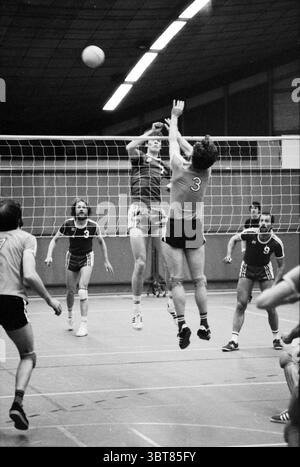 Phoenix - VCIJ Volleyball, Whizgle News, Dutch Desk, Niederlande, 1950 - 2000 auf 31-03-1979. Die Abbildung zeigt diese Themen. Die Szene fängt einen intensiven Moment während eines Volleyballspiels in einem geräumigen Hallenplatz ein. Auf der einen Seite des Netzes springt ein großer Spieler in die Luft, sein Körper wurde vollständig ausgestreckt, während er sich auf ein dramatisches Spiel vorbereitet. Er trägt ein Trikot mit auffälligen Streifen und wirkt fokussiert, mit einem entschlossenen Ausdruck. Die Maßnahme schlägt hohe Energie und Wettbewerbsfähigkeit vor. Ihm gegenübersteht ein anderer Spieler, der leicht gehockt und ebenfalls bereit ist zu springen, die Nummer 3 auf seinem Trikot Stockfoto
