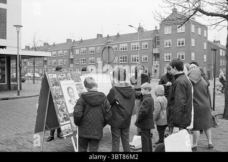 Wall Newspaper Market Square Miscellaneous, Whizgle News, Dutch Desk, Niederlande, 1950 - 2000 am 01-03-1969. Dies sind die Elemente im Bild. Die Szene zeigt eine Gruppe von Personen, die sich um eine Schautafel versammelt haben, auf der verschiedene Poster und Kunstwerke gezeigt werden. Die Umgebung hat ein urbanes Flair, gekennzeichnet durch flache Gebäude mit einheitlichem Architekturstil, die eine Mischung aus Ziegelstein und Betonoberflächen bieten. Diese in gedämpften Tönen gemalten Strukturen schaffen eine etwas melancholische Atmosphäre. Die Einzelpersonen führen Gespräche, was auf eine gemeinschaftliche Aktivität oder Veranstaltung hinweist. Darunter Stockfoto