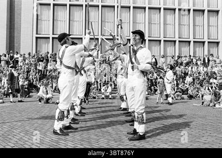 Morris-Tänzer in IJm. Dancing, Whizgle News, Dutch Desk, Niederlande, 1950 - 2000 am 03-09-1977. Dies sind die Elemente im Bild. Die Szene fängt ein lebhaftes Outdoor-Event voller Aktivität ein. Im Vordergrund steht eine Gruppe von Männern in traditioneller weißer Kleidung, die jeweils schwarze Hüte tragen und mit bunten Schärpen über ihren Körper hängen. Sie spielen eine synchronisierte Performance, jonglieren geschickt und werfen verschiedene Objekte, wahrscheinlich Stäbe oder Schlagstöcke, hoch in die Luft. Die Gesichtsausdrücke vermitteln eine Mischung aus Konzentration und Freude, Showcasin Stockfoto